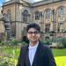  Krish Wadhwani is wearing his graduation gown with a white shirt and blue tie, with short back hair with glasses looking standing in front of a classic building.