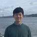 Kun Lee, with short black hair and wearing a green top, smiles at the camera. A bridge and a body of water are in the background.
