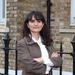 Merve is smiling and standing with her arms folded in front of a brown brick building. She has long dark hair and a fringe. She is wearing a white shirt and brown blazer. 