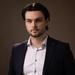 Matej has medium-length dark hair. He is wearing a white shirt and dark blazer. 