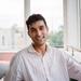 Karan Kalsi has short dark hair and is wearing a loose striped shirt. He is sitting in front of some windows and smiling at the camera.