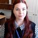 Kayleigh is sitting down with long red hair, wearing a dark blue vest and checked blue and white shirt, looking right at the camera.