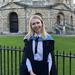 Woman with blond hair, wearing university gown standing by a metal rail with University building in background