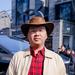 Yang Jianing is wearing a brown cowboy hat, cream jacket and red and black striped shirt with glasses looking at the camera.