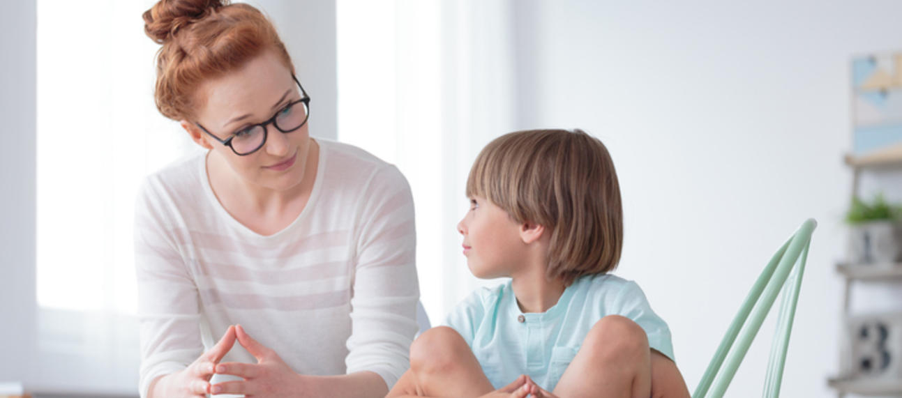 A young boy sits cross-legged on a chair, looking up at an adult woman sat next him.