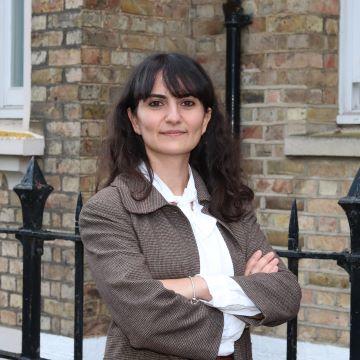 Merve is smiling and standing with her arms folded in front of a brown brick building. She has long dark hair and a fringe. She is wearing a white shirt and brown blazer. 