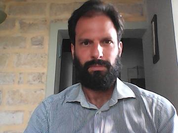 Paolo Graziano wears a blue striped shirt, has a beard, and is sitting in front of a brick wall