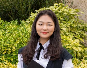 Jinseo Kim is wearing graduation gown, with dark shoulder length hair smiling at the camera.