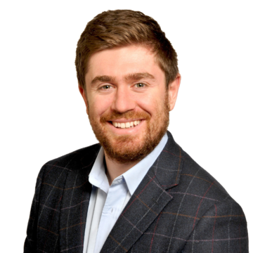 Benjamin Scher has short ginger hair and a beard, wearing a blue shirt with a dark blue suit jacket, and smiling at the camera.