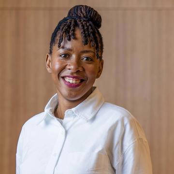 Dr Nontokozo Langwenya wears a white shirt and is smiling in front of a wood-panelled background.