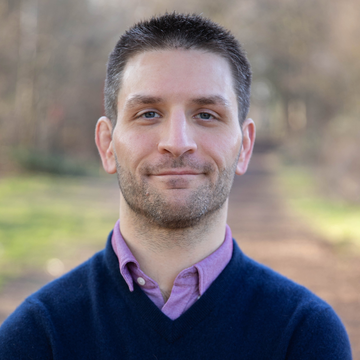 Lewis Anderson is wearing a dark blue sweater over a purple shirt, with short dark hair, looking directly at the camera.