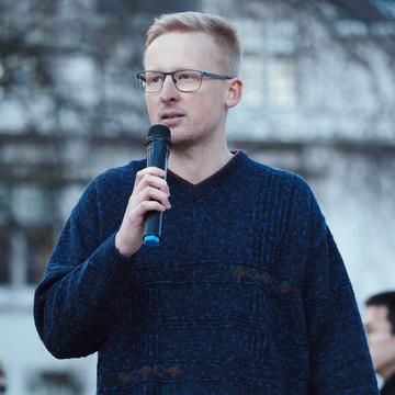 Benjamin is standing outside, wearing a navy jumper and speaking into a microphone.