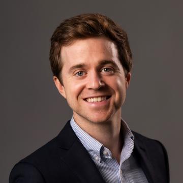 Zach Parolin has short brown hair. He is wearing a light blue shirt and a dark blazer, and smiling broadly. 