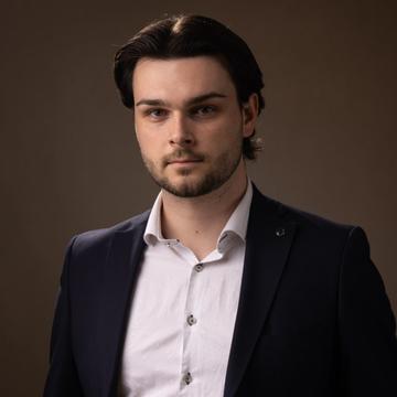 Matej has medium-length dark hair. He is wearing a white shirt and dark blazer. 