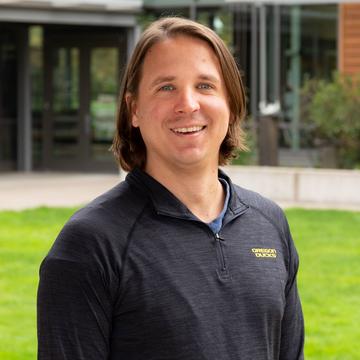 Sean has long straight brown hair and blue eyes. He is standing outside on some grass, wearing a dark grey lightweight sport jumper and smiling at the camera.
