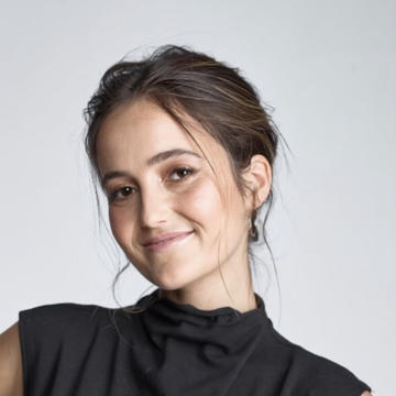 Eliana has brown hair in an updo and is wearing a high-neck draped black top. She is smiling at the camera.
