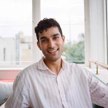 Karan Kalsi has short dark hair and is wearing a loose striped shirt. He is sitting in front of some windows and smiling at the camera.