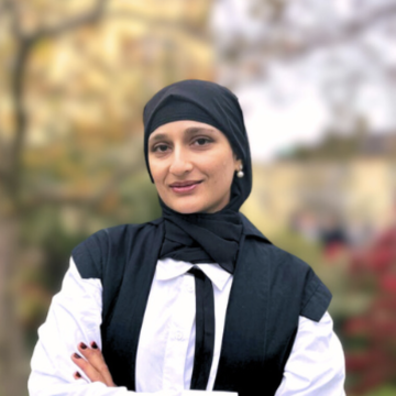 Kayanat Iltaf stands with folded arms wearing academic dress in front of an out-of-focus tree in the background. 