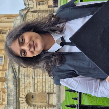 Shrutika is standing in front of the Radcliffe Camera in Oxford. She is wearing subfusc clothing, including a white shirt, black gown, black tie and black cap.