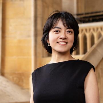 Wearing a black dress and pearl drop earrings with a short hair cut, Yi Qie smiles at the camera.